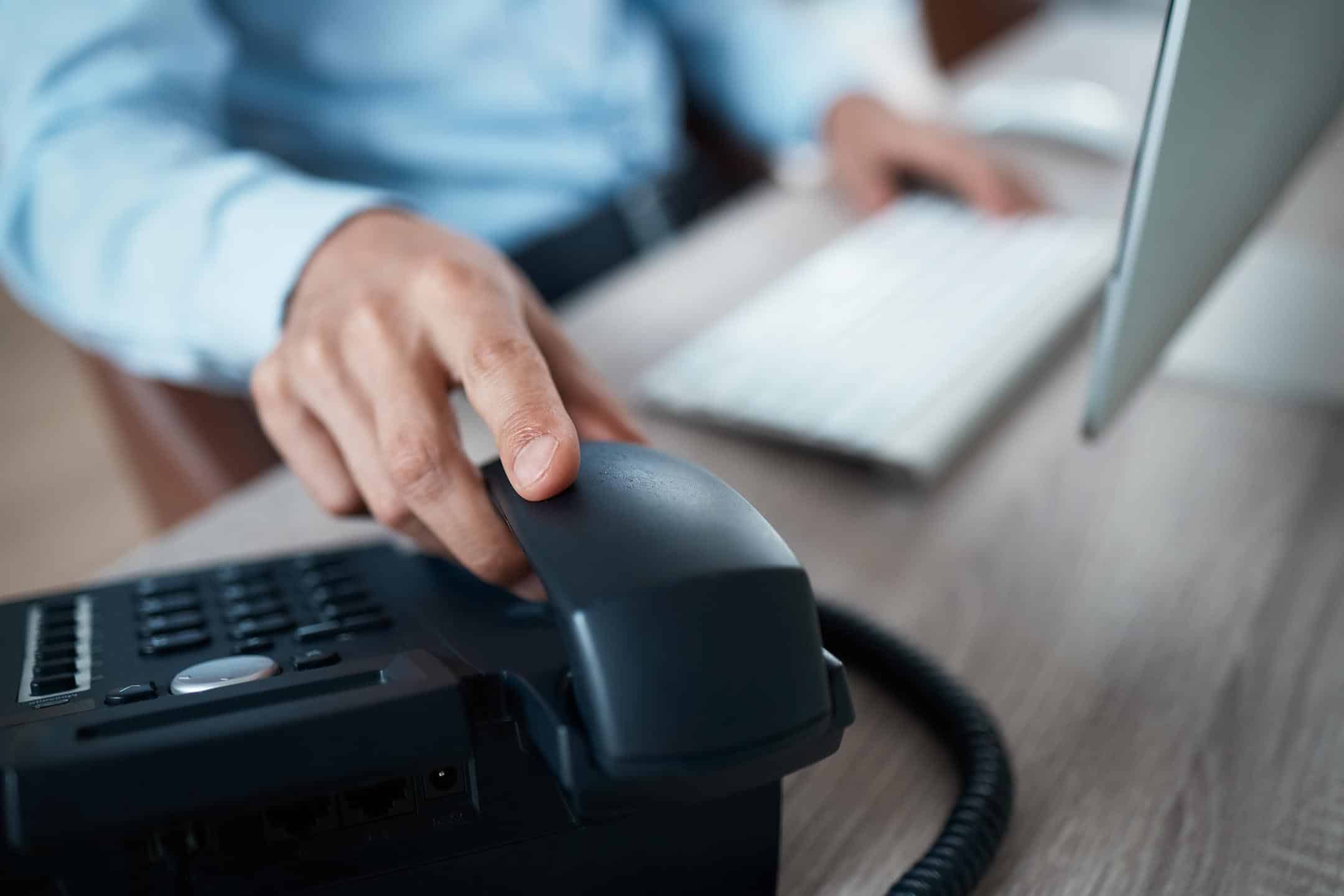 Person using a landline phone with a computer in the background.