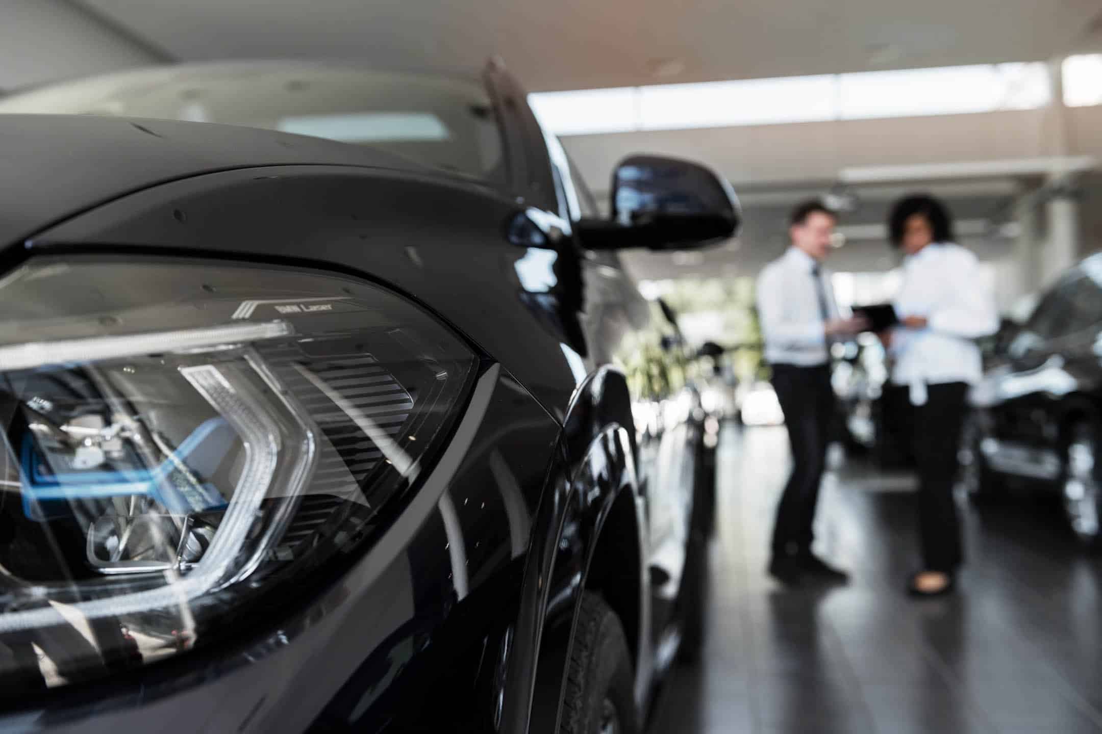 Close-up of a black luxury car in a showroom with two people in the background.