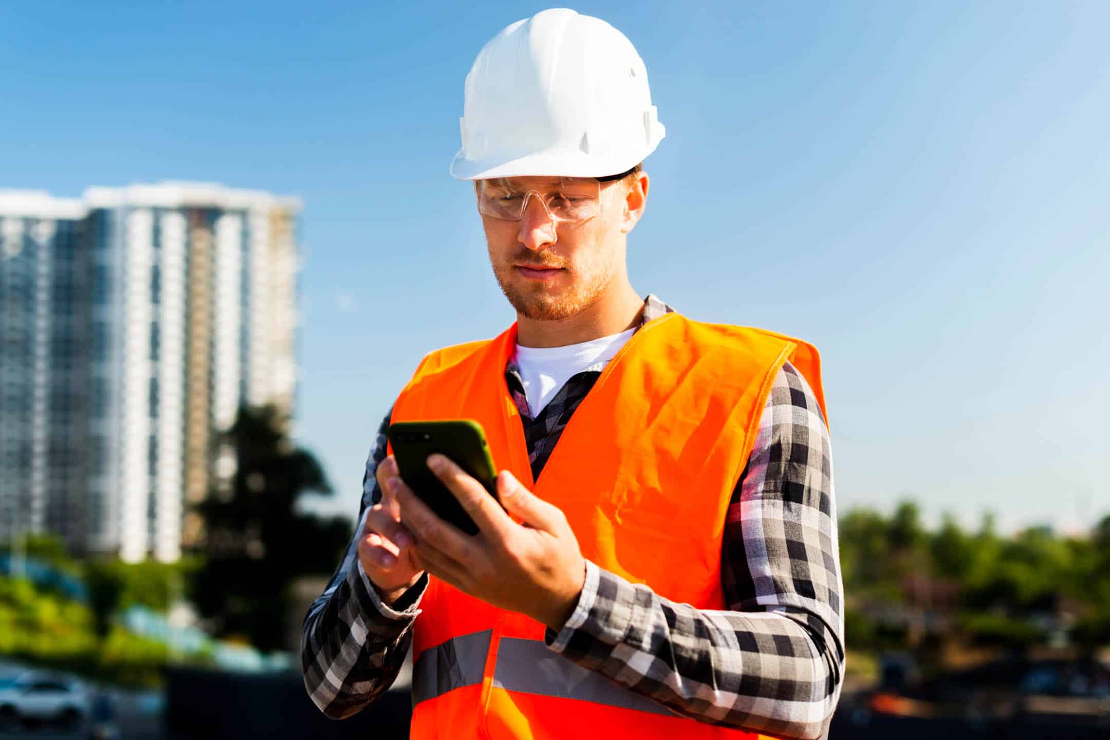 Construction worker in safety gear using smartphone outdoors.