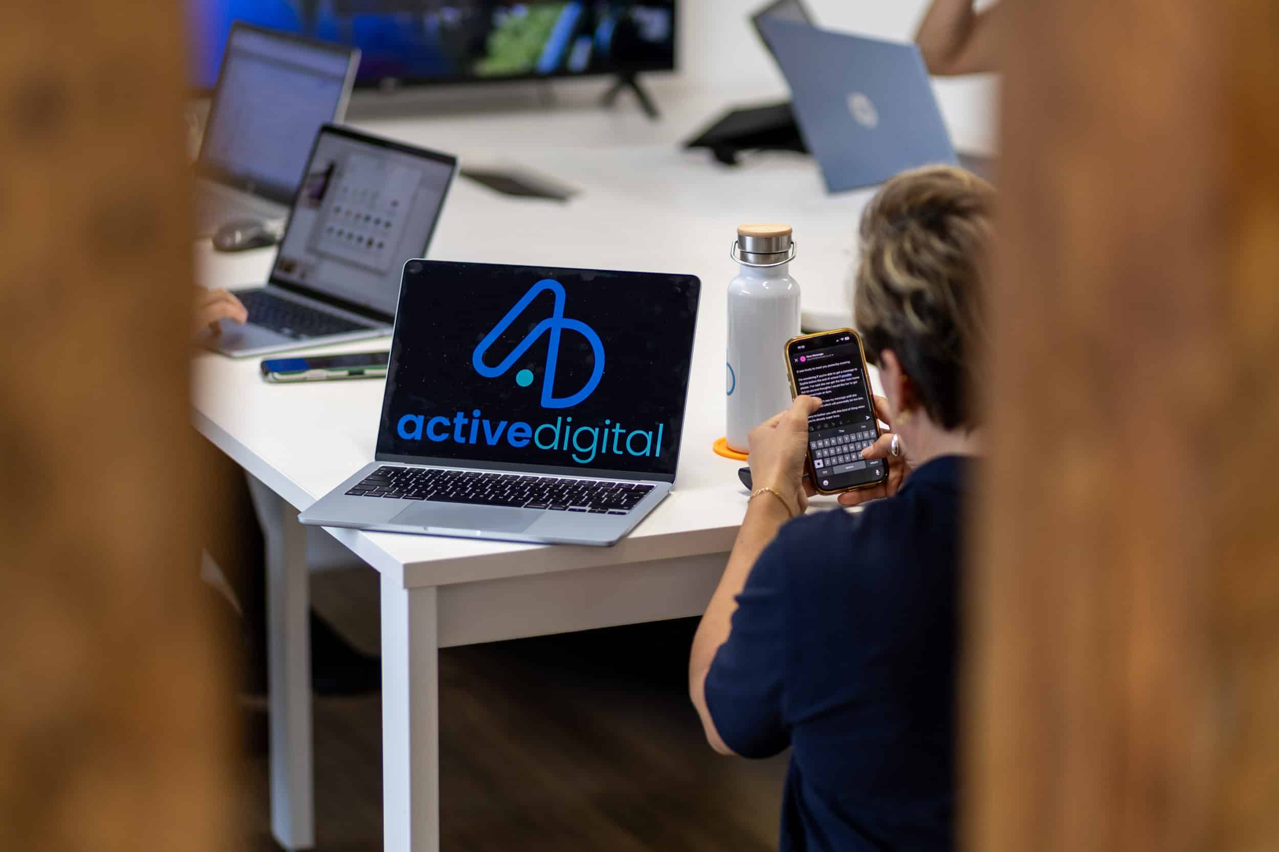 Person using smartphone in a modern office with laptops and digital devices.